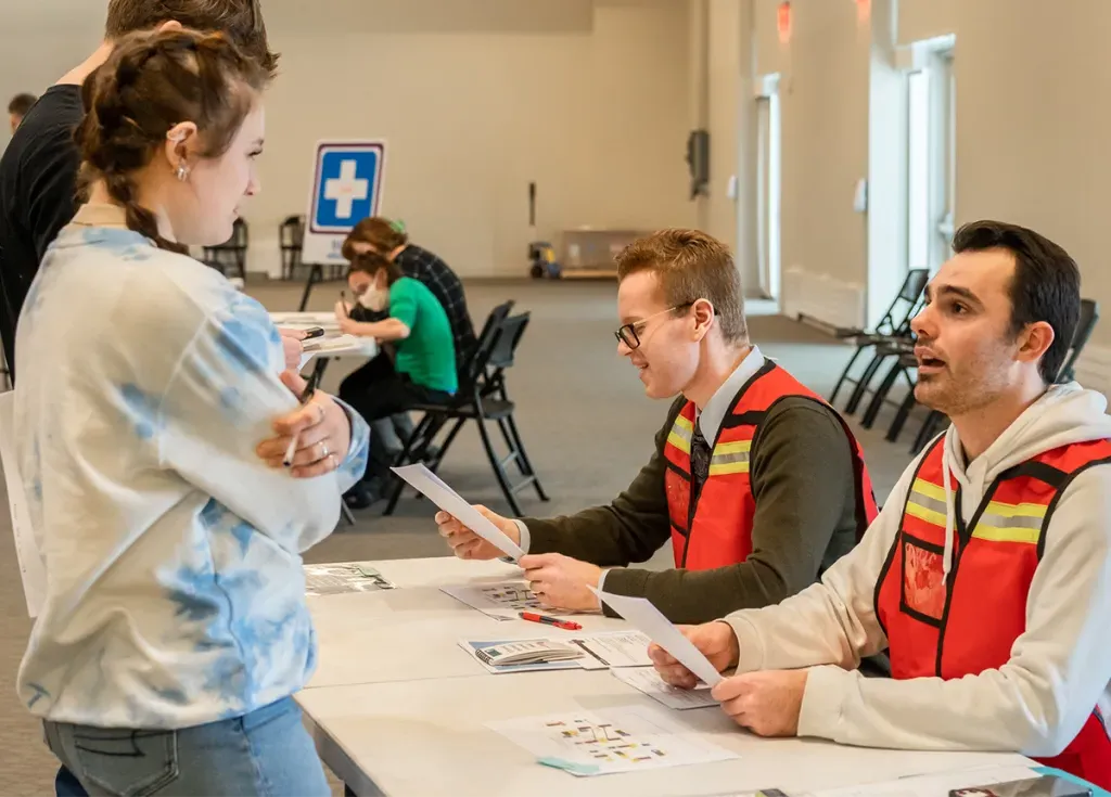 Two students in orange vests talk to a small crowd at a public health event